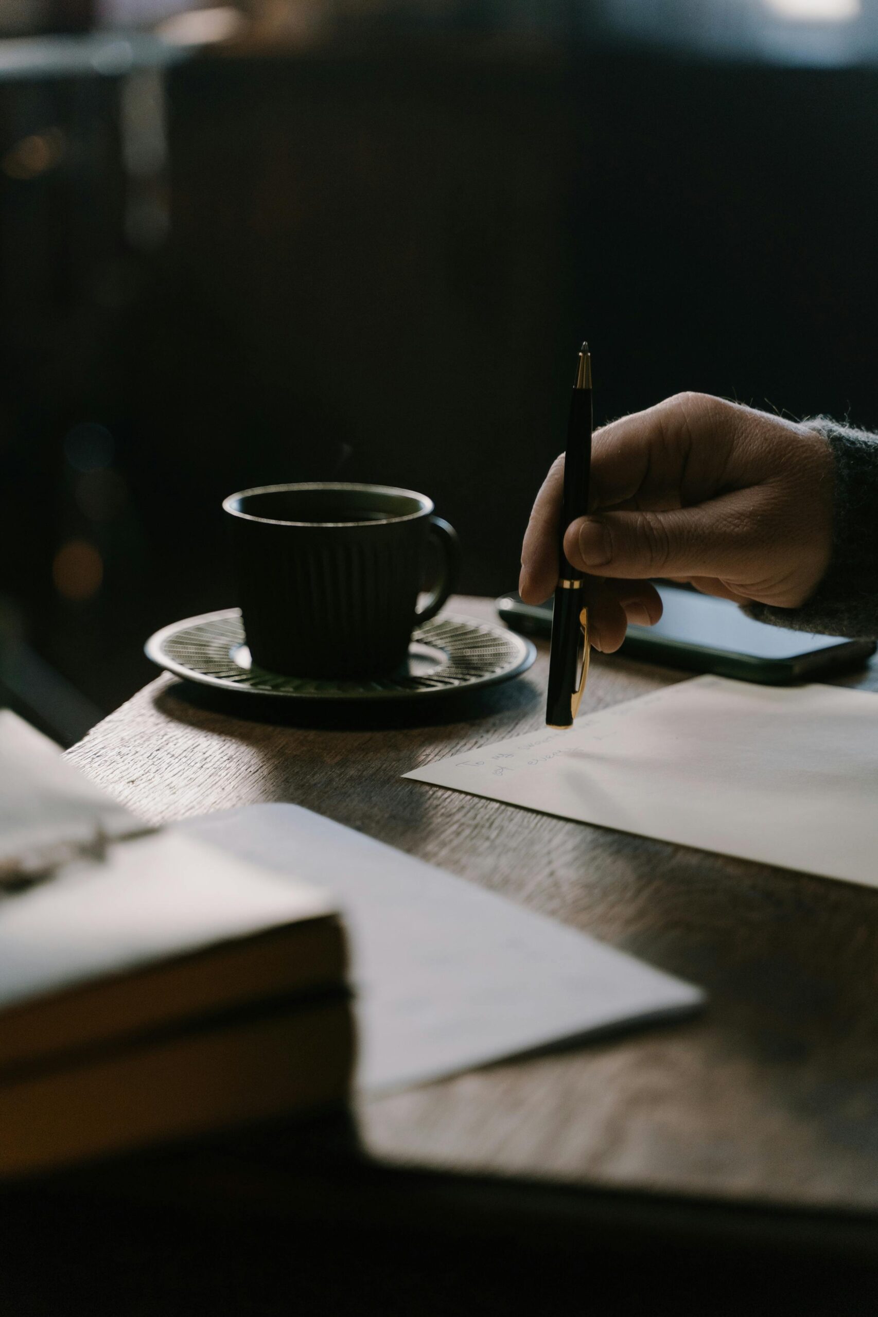 A warm, moody setting featuring a hand holding a pen, coffee cup, and journal on a wooden desk.